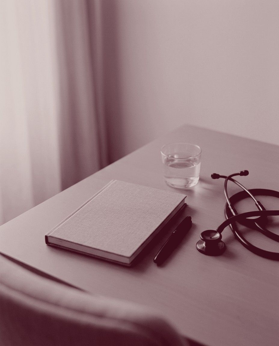 Mesa de consultório com estetoscópio, caderno, caneta e um copo de água, foto em tons de marsala